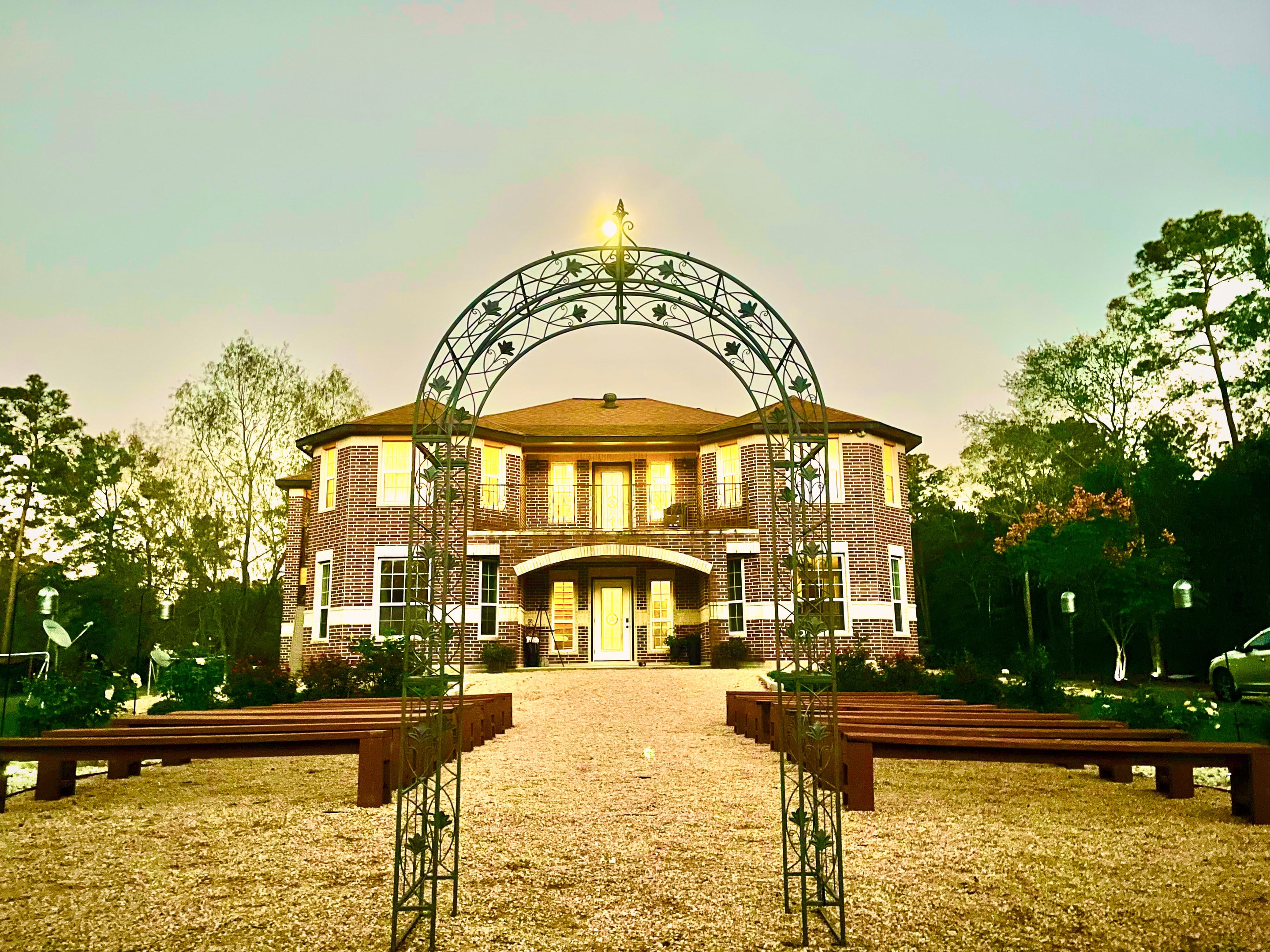 The brick manor house at dusk, framed by a wrought-iron arch, its windows lit