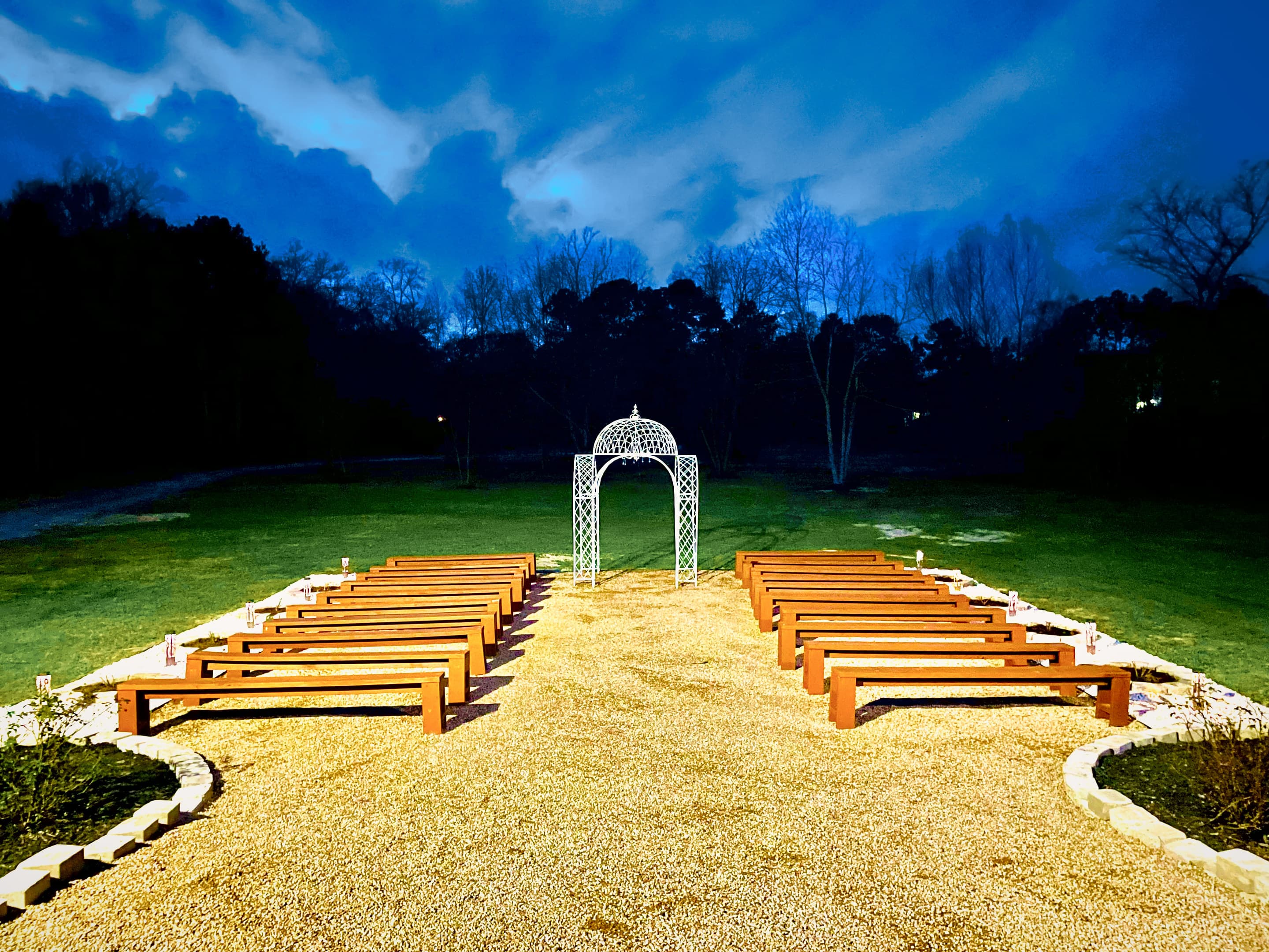 The garden ceremony aisle at blue hour, benches softly lit beneath a darkening sky