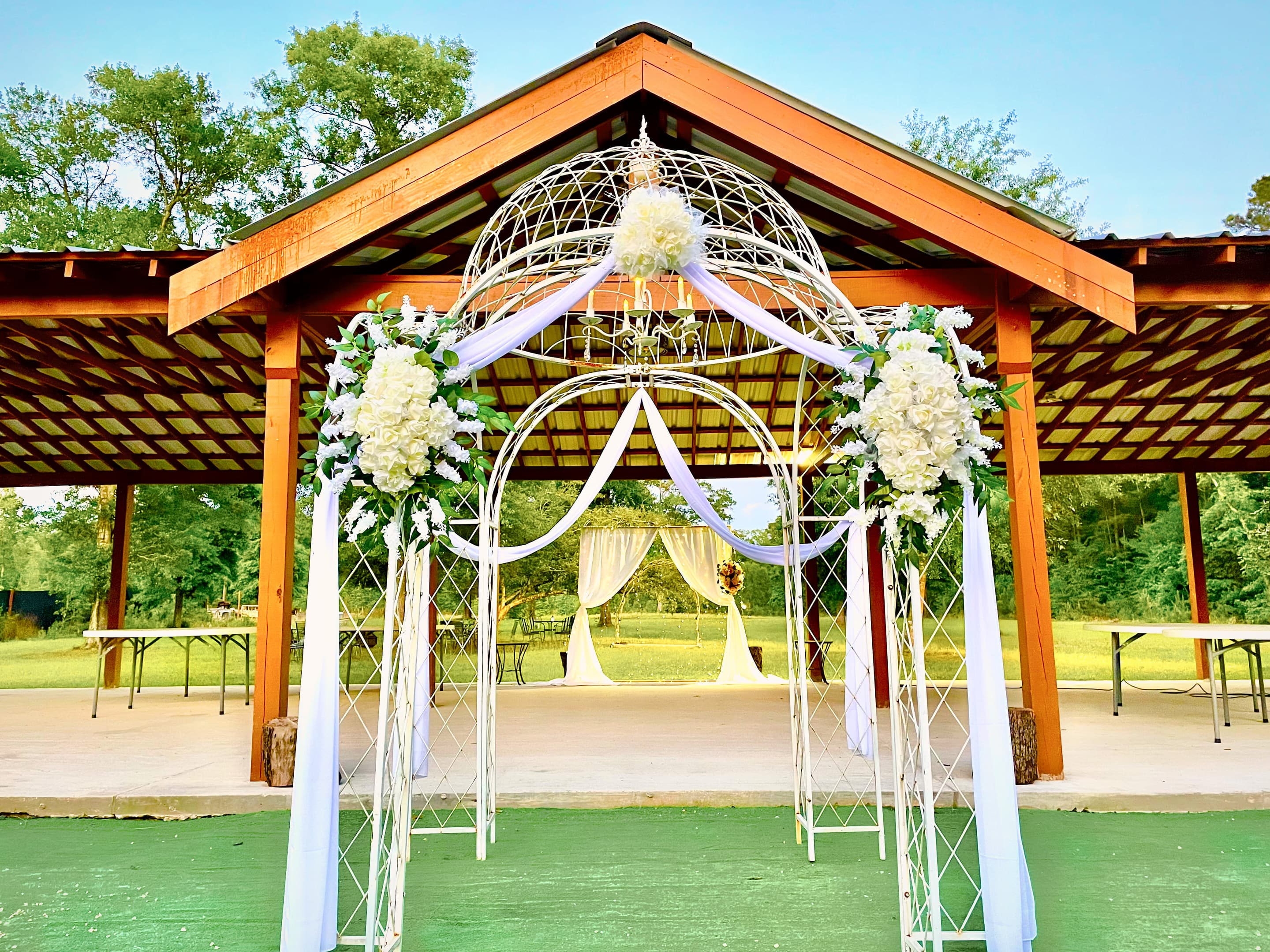 The pavilion prepared for a ceremony, its wrought-iron arch dressed with white florals